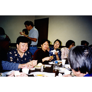 Women sit and eat Thanksgiving dinner, with a man and two women carrying their plates in the background