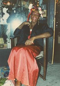A Photograph of Marsha P. Johnson Sitting Wearing a Floral Headpiece, Blue Jacket, and Red Dress