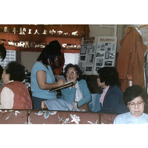 Woman wearing a blue short-sleeve sweater writes on a notebook while a woman wearing a blue cardigan looks on