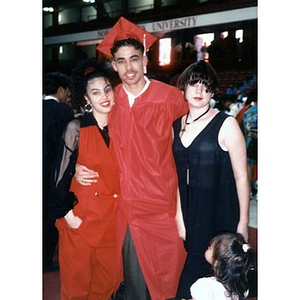 Young man in a red graduation cap and gown stands with two young women in front of a Northeastern University building.