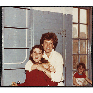A woman posing for the camera with her arms around a teenage girl while a younger girl sits at the window behind them at the Boys and Girls Club