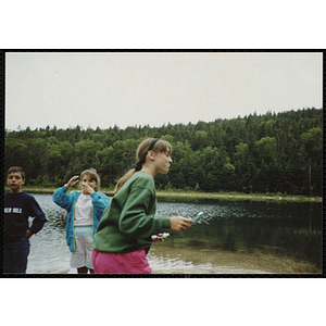 A girl brushes her teeth on the shore of a lake as a girl and boy look on