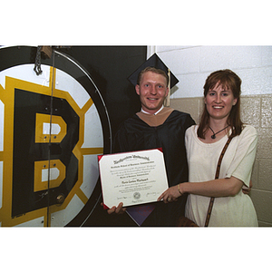 Standing by a Boston Bruins banner in his academic robes, Nevin Markwart of the Boston Bruins holding his diploma poses with a woman