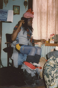 A Photograph of Marsha P. Johnson Sitting in a Chair Wearing a Pink Hat, Blue Vest, and Blue Tights
