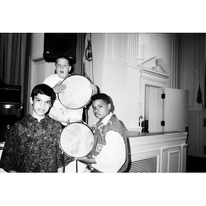 Three boys pose by the edge of the stage holding tambourines.