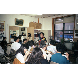 Association members sit and eat, listening to a woman in front of them