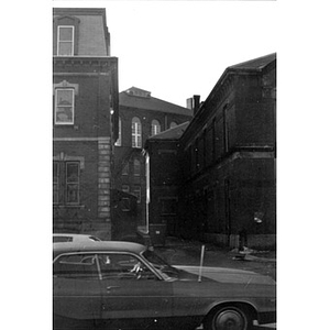 View of the alleyway between La Alianza Hispana's headquarters, 409 Dudley Street (building on left) and 407 Dudley Street (on right), taken from across the street; there is a parked car and another car driving by in the foreground