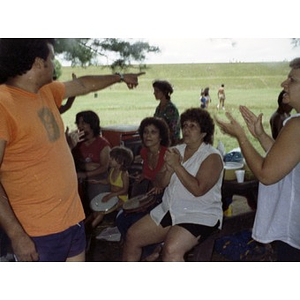 People seated and standing by a picnic table clap their hands and play musical instruments at a La Alianza staff picnic, while the man on the far left points to something in the open field behind them