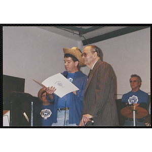 A Bunker Hillbilly alumnus holds a microphone and reads from a document with an unidentified elderly man at his side