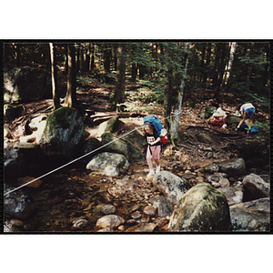 A girl with a backpack crosses a creek holding a rope line on the Piper Trail