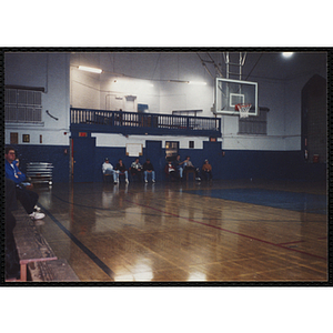 A group of teenage boys sit behind the basket in chairs as they watch the action on the other half of the court during a basketball game