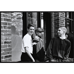 Three teenage boys standing together, while one of them looks at the camera, in front of a window outside the Charlestown Boys & Girls Club building