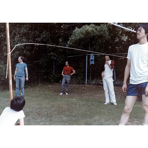 Male Association members stand near a volleyball net and look to their right, while a young boy crouches near the net and watches them