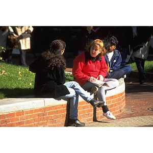 Two students converse while sitting on the stone wall at the edge of the library courtyard