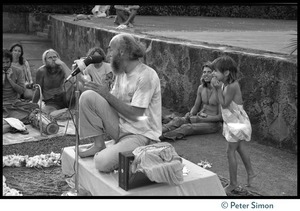 Ram Dass seated on a small platform during his appearance at Andrews Amphitheater, University of Hawaii, a small child sneaking up behind