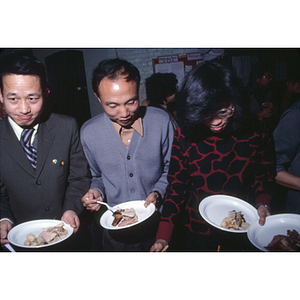 Two men and one woman stand in a row and serve themselves food at a buffet table