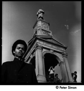Unidentified African American man standing by the Civil War memorial in Cambridge Common, antiwar protesters clambering over the memorial