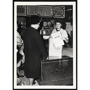 A woman stands behind the counter in a grocery store and looks at the camera with a flyer displaying the text "Members of the BOYS' CLUBS OF BOSTON" and "VOTE" while the customers look on