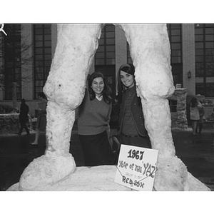 Two students stand between the legs of a giant snow sculpture at the Winter Carnival