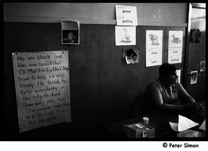 Teacher seated at a table at the Liberation School: in front of a poster honoring Martin Luther King: 'He tried to help everbody in the city. He died. Somebody shot him...'