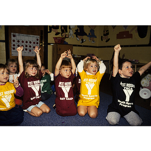 Children wearing West Roxbury-Roslindale YMCA shirts seated on a classroom floor, with their arms raised