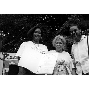 Two women and a man on the outdoor stage at Festival Betances displaying a certificate.