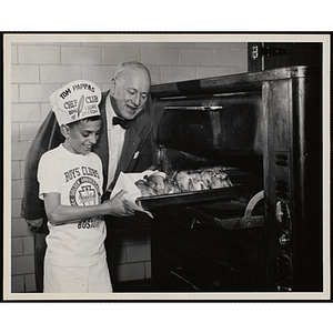 A member of the Tom Pappas Chefs' Club removes a pan of cooked chickens from an oven in a Brandeis University kitchen as Executive Director of Boys' Club of Boston Arthur T. Burger looks on