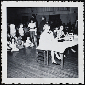 The panel of judges sit at a table while the audience sits and stands behind them during a Boys' Club Little Sister Contest