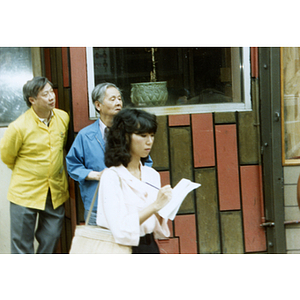 Two Asian men and one Asian woman standing outside a storefront in Chinatown watching demonstrators march through the street demanding justice for Long Guang Huang