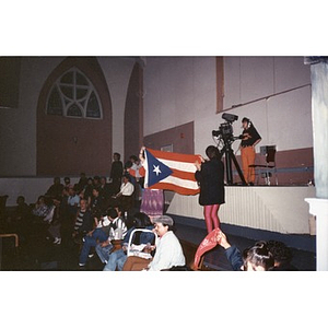 Two women display a large Puerto Rican flag from the back of a school auditorium.