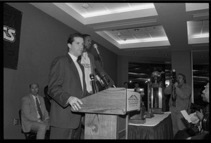 John Calipari at the microphone during a press conference with Marcus Camby (background)