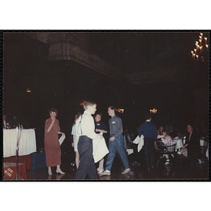 A boy holding a white bag and walking past the camera while the guests sit and stand near the head table during the "Recognition Dinner at Harvard Club"