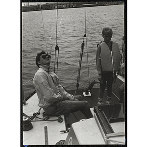 A boy stands next to a seated man on the deck of a sailboat in Boston Harbor