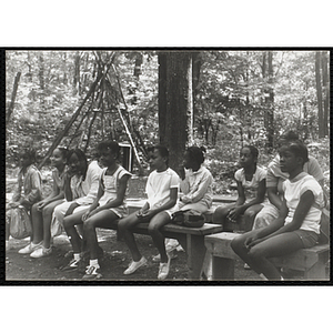 A Group of girls sitting on benches in a wooded clearing at camp