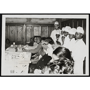 A group of boys dressed as chefs stand behind people seated at a table during a Moms' Club event