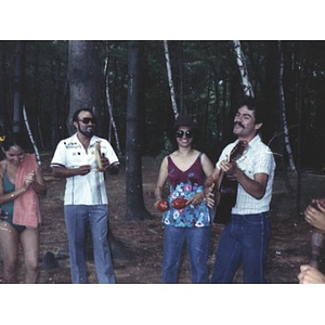 Group of four people stand in the woods playing musical instruments, singing, or clapping their hands at a La Alianza staff picnic at an unidentified location