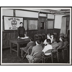 A man stands at a table and addresses a group of seated boys