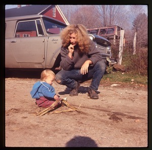 Charles Light and baby Eben crouching by a car (Scout), in front of the house at Montague Farm Commune