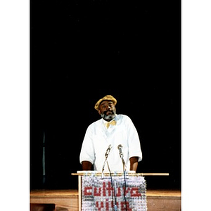 Man in a yellow hat and bow tie at the podium during a Cultura Viva event.