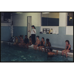 A woman speaks to a group of children sitting on the edge of a natorium pool