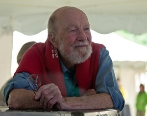 Pete Seeger: portrait seated at a table with a red sweater around his neck at the Clearwater Festival