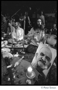 Ram Dass seated on stage (left of center) while Krishna Das sings, with portrait of Neem Karoli Baba in foreground