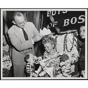 A man places a crown on the Little Sister Contest winner as she sits in a decorative chair with a doll