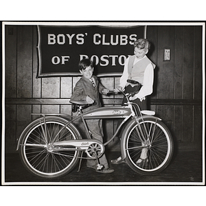 A boy, wearing a suit, posing with a bicycle in front of a Boys' Clubs of Boston banner while a woman stands beside him
