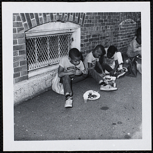 Three boys sit against a wall and eat at a picnic