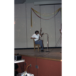 Man seated on stage with instrument during Chinese Progressive Association's Fifth Anniversary Celebration