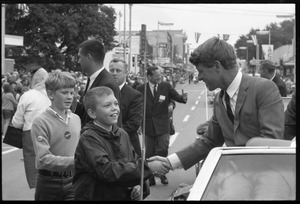 Robert F. Kennedy (left) seated in an open car at the Turkey Day parade, shaking hands with two boys; while stumping for Democratic candidates in the northern Midwest