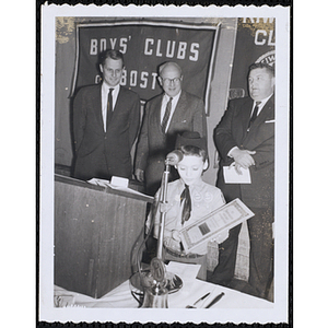 A boy wearing a cowboy hat and a handkerchief necktie displays his certificate at the podium during a Kiwanis Club's awards ceremony