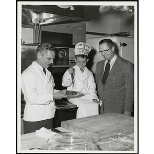 A seated member of the Tom Pappas Chefs' Club holding a fork over a plate of meat poses with two men in a kitchen