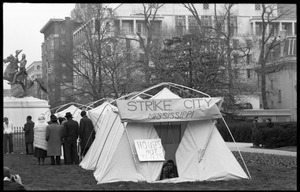 Man lying his tent, looking out the front flap: signs reading 'Strike City Mississippi' and 'Houses not tents,' statue of Lafayette in the background